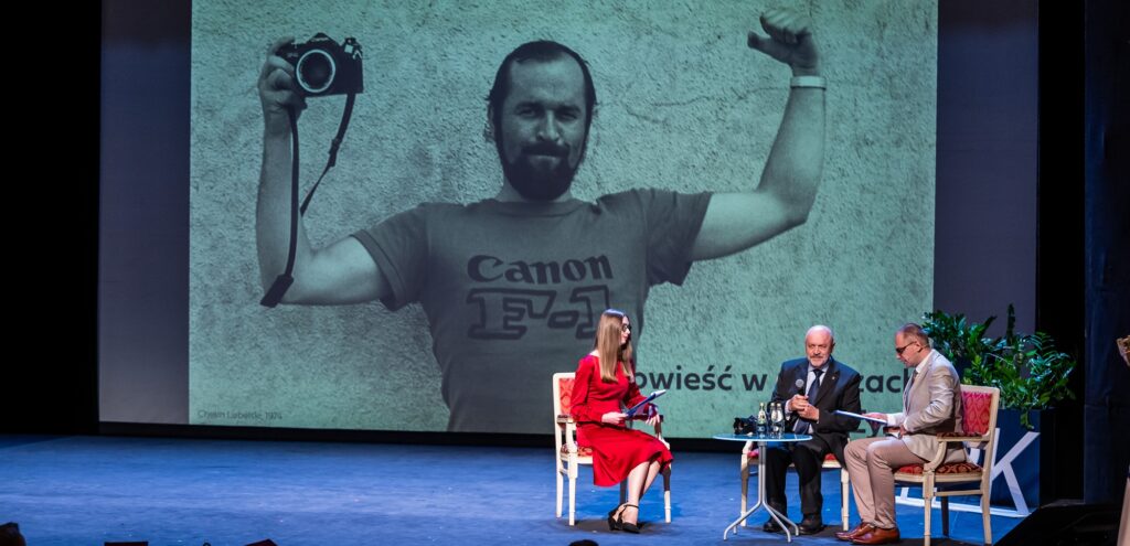 Jan Zych na scenie Teatru im. Juliusza Słowackiego odpowiadał na pytania Wiktorii Duś i Bartłomieja Krystyńskiego, w tle zdjęcie sprzed pół wieku, otwierające wystawę „Opowieść w obrazach”. Fot.: Piotr Malec